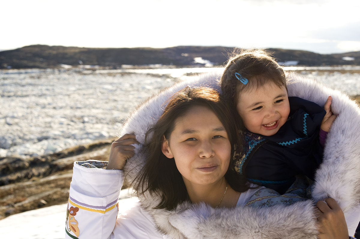An Inuit mother has her daughter in her amouti, a traditional way to carry the young people.  This is on the tundra of Baffin Island, Nunavut, Canada in late spring. Smiling.  Copy space.